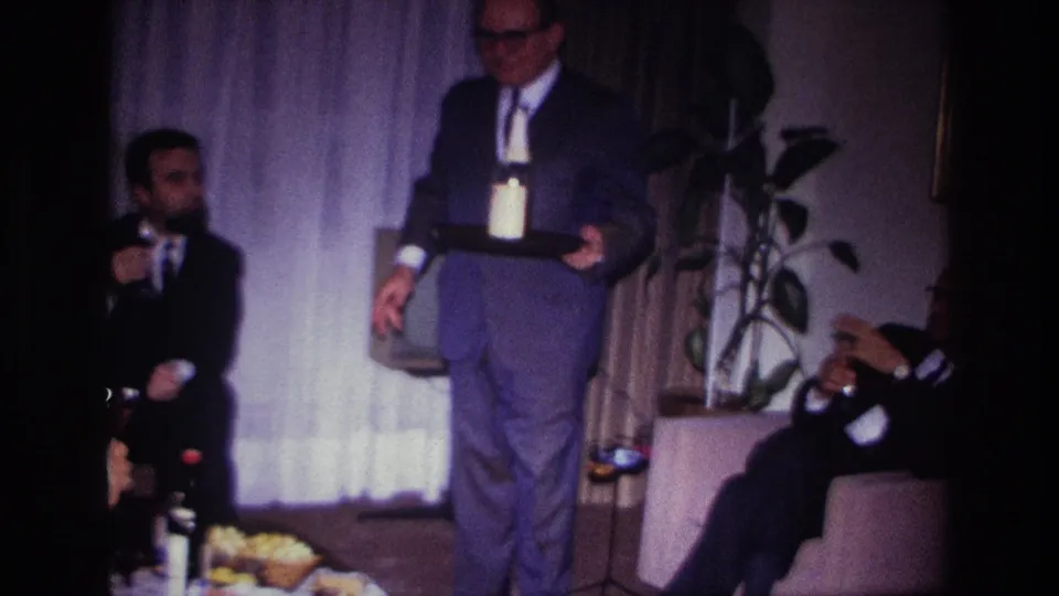 1967 Rockaway Beach living room frame showing a man serving drinks as a seated guest gestures beside curtains and a potted plant.