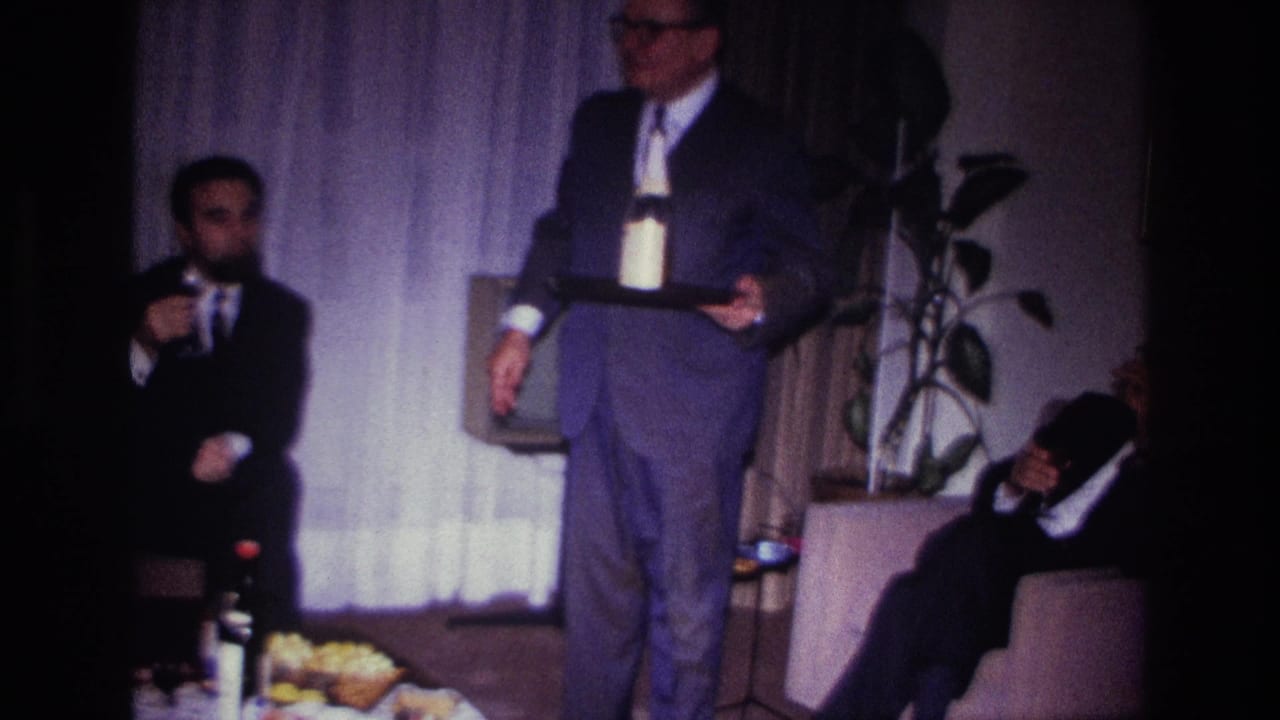 1967 Rockaway Beach home-movie frame showing a suited man holding a tray with a bottle and glass as guests sit in a private living room.