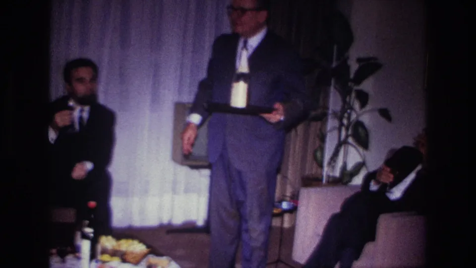 1967 Rockaway Beach home-movie frame showing a suited man holding a tray with a bottle and glass as guests sit in a private living room.