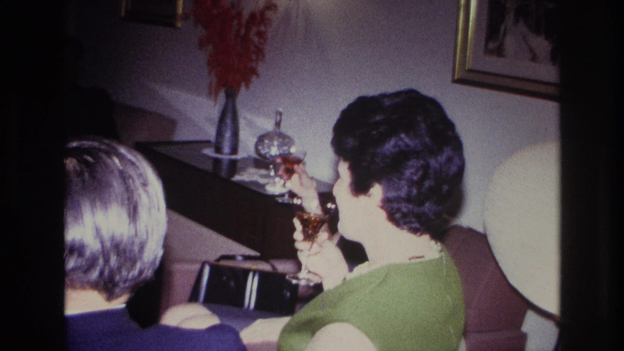 1967 Rockaway Beach home-movie frame with a woman in a green dress holding a cocktail glass beside another seated guest in a private interior.
