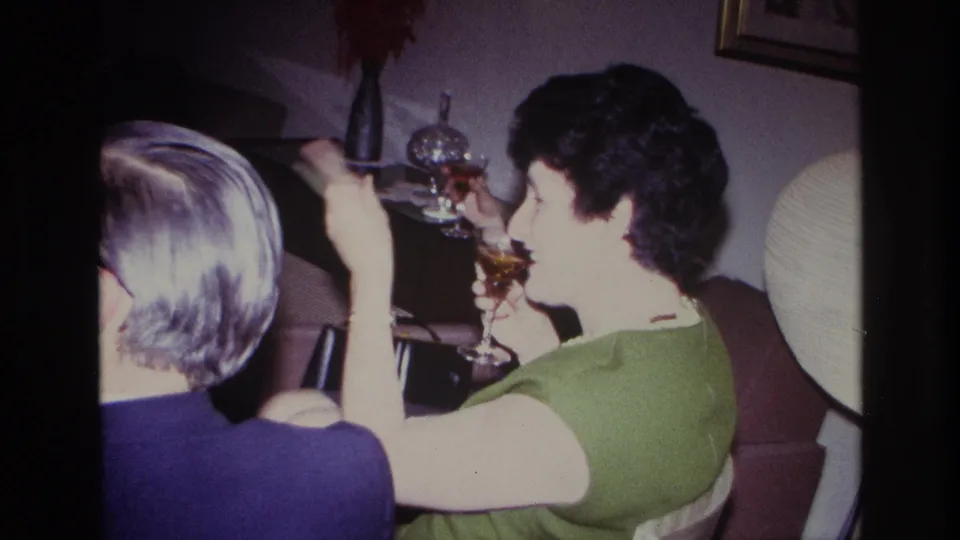 1967 Rockaway Beach home-movie frame showing seated adult guests with drinks in a warmly lit private room with mid-century decor.