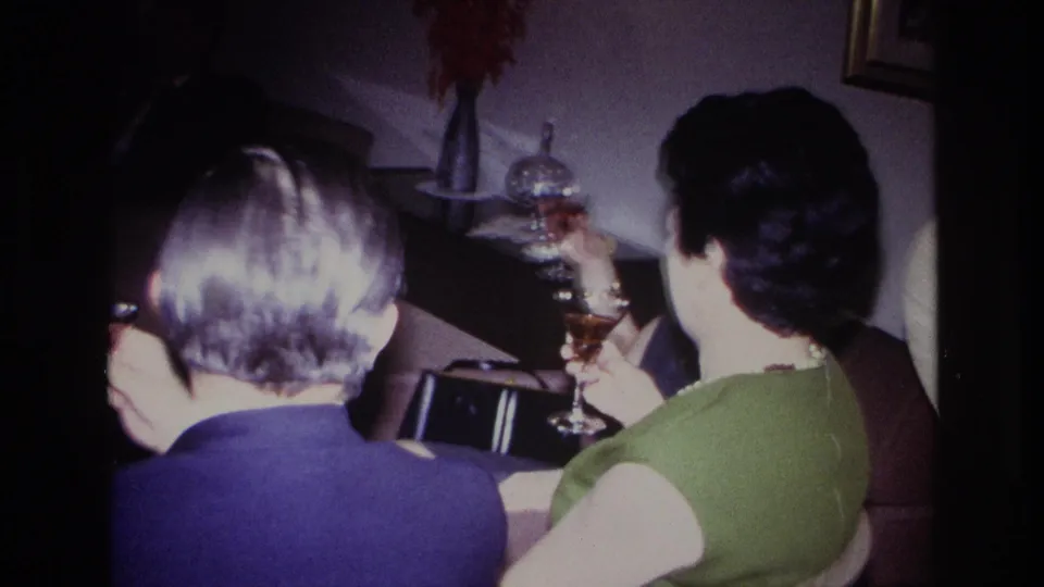 1967 Rockaway Beach home-movie frame of seated party guests from behind, with a woman in green holding a glass in a private living room.