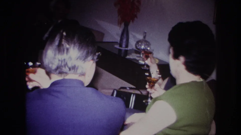 1967 Rockaway Beach home-movie frame of adults raising cocktail glasses in a dim private living room.
