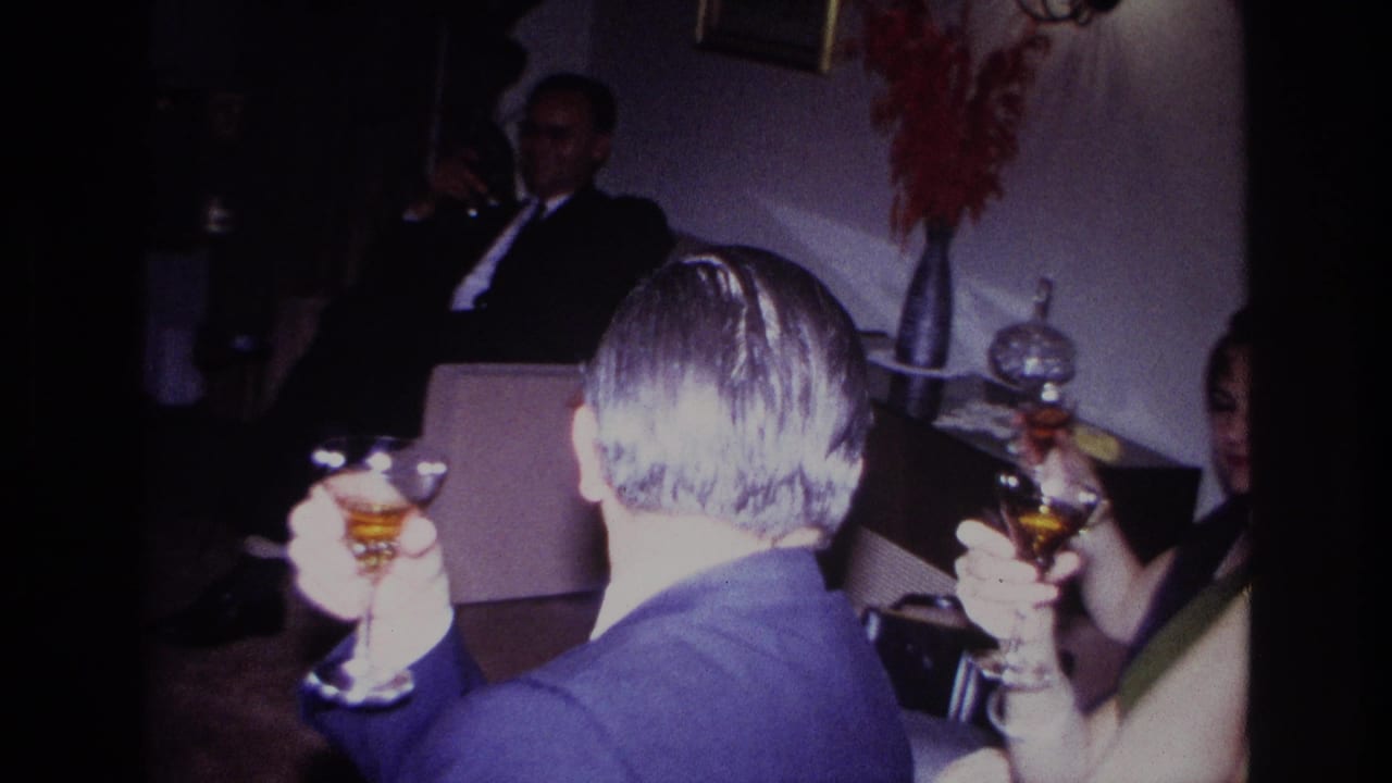 A man in glasses and nearby adults hold drinks in a living room with a couch behind them.