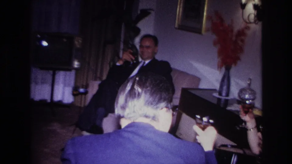 Adults with drinks gather in a Rockaway Beach living room in 1967, with a suited man on a couch and a small TV nearby.