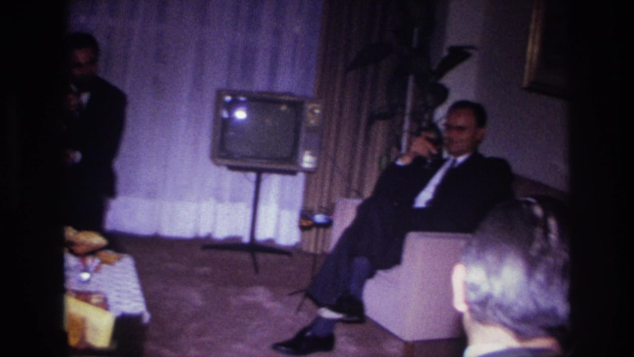 A suited man sits with a drink beside a small television in a softly lit living room.