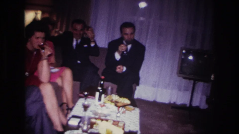A woman in a red top sips a drink near a table as suited men sit behind her in the Rockaway Beach home-movie scene.