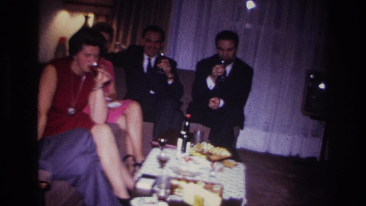 1967 Rockaway Beach home-movie frame showing adults seated indoors with wine glasses, bottles, and snacks on a low table.