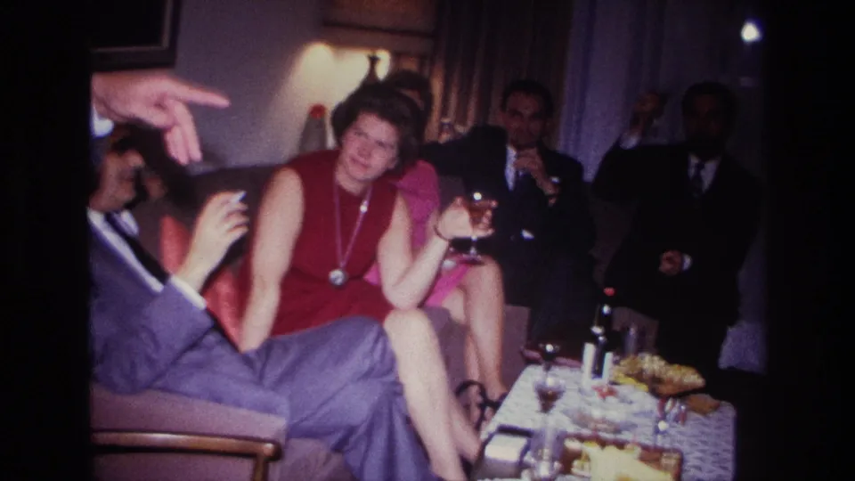 1967 Rockaway Beach home-movie frame of adults conversing, with a man gesturing and a woman smiling with a glass.