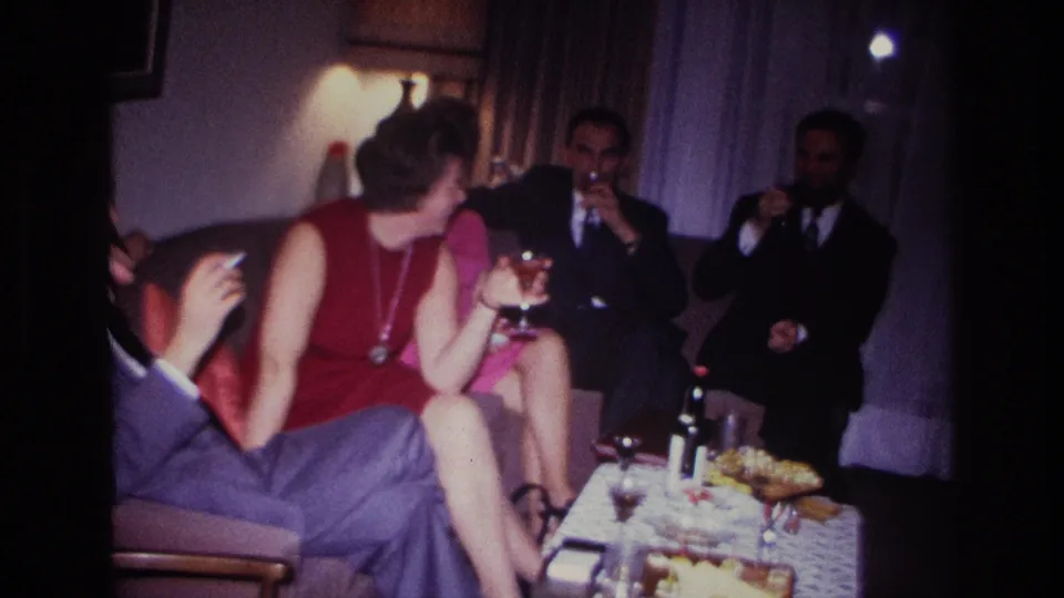 1967 Rockaway Beach indoor gathering where a woman turns toward suited men as drinks and snacks sit in the foreground.