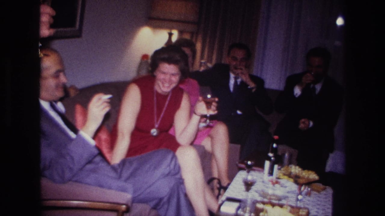 1967 Rockaway Beach home-movie frame of adults socializing indoors, including a laughing woman in red and a man in a blue suit smoking.