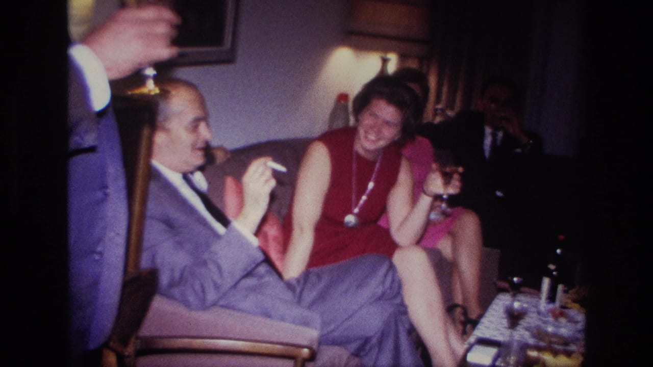 Seated adults converse in a living room while holding drinks, with a lamp and table visible nearby.