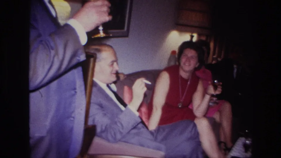 1967 Rockaway Beach home-movie frame with a standing guest in a suit, seated adults, drinks, and warm living-room lamp light.