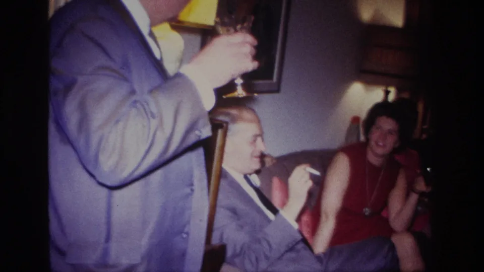 1967 Rockaway Beach frame with a foreground suited guest holding a glass and seated adults socializing in a private living room.