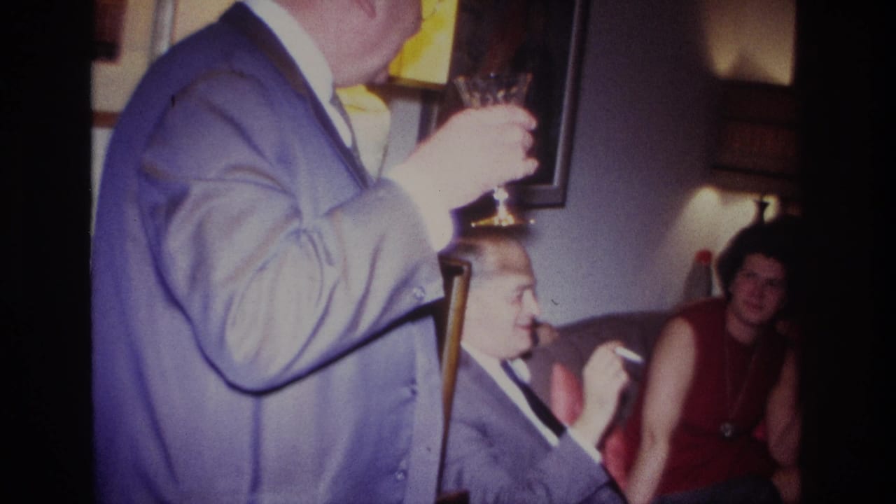 In a 1967 Rockaway Beach home movie frame, a man in a blue suit holds wine as adults sit nearby in a warmly lit living room.