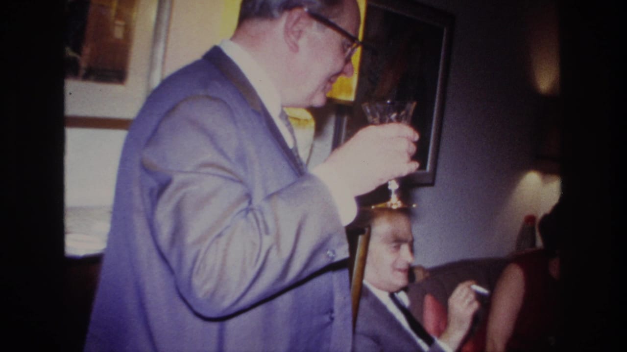 Man in glasses holds a wine glass near his face as seated guests remain partly visible.