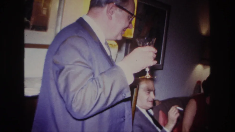 A suited man with glasses holds a wine glass close to his face in a private Rockaway Beach living room, with other adults partly visible.