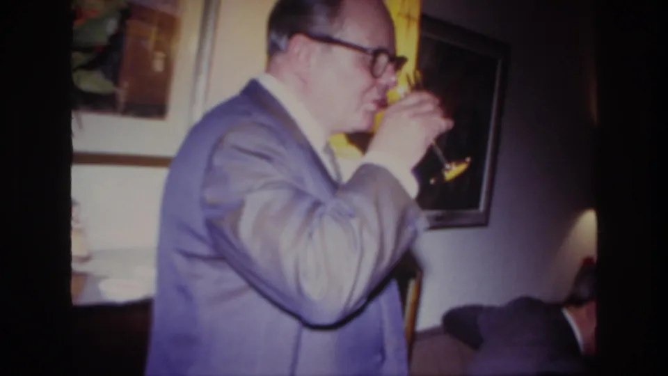 The home-movie frame shows the suited man sipping wine in a warm interior, with framed wall art and a couch edge visible.