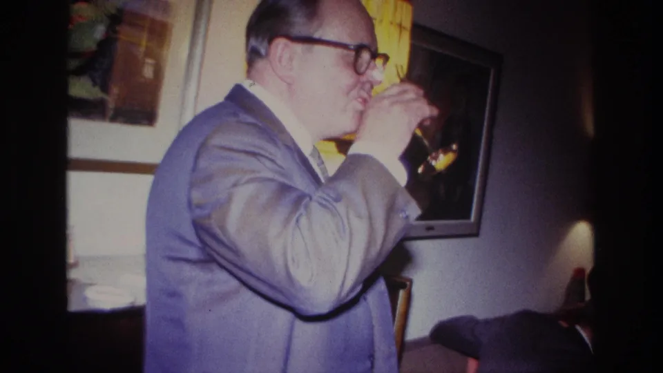 A 1967 Rockaway Beach living-room frame shows the suited man drinking wine, with another seated adult partly visible at the edge.