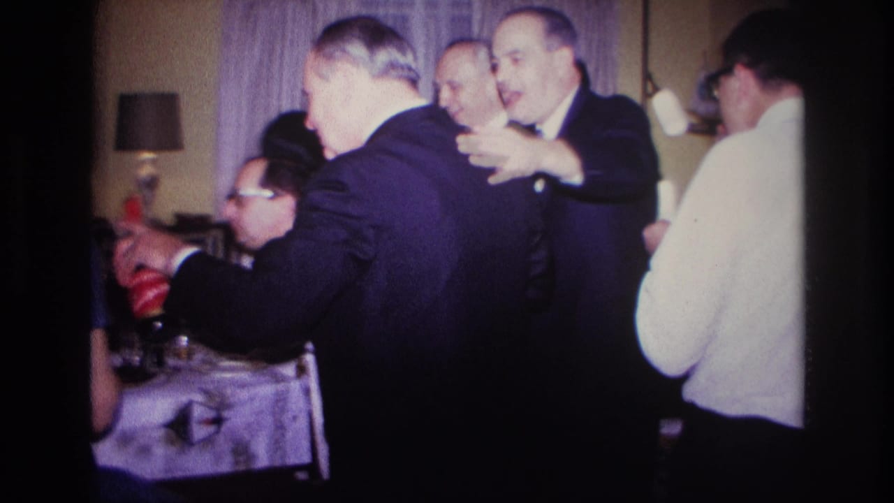 1967 Rockaway Beach home-movie frame showing suited men gathered indoors around a table during a lively private celebration.