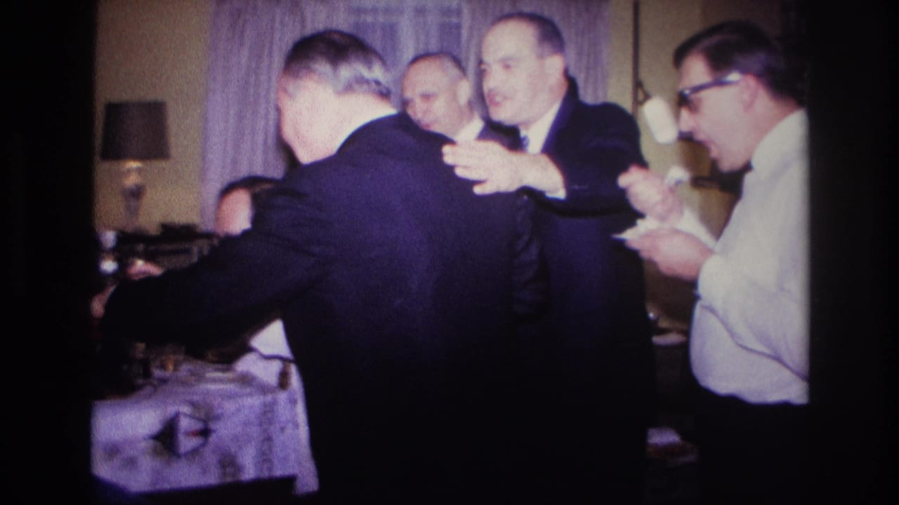 A suited man faces away while other men smile, gesture, and hold small plates indoors.