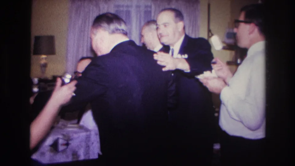1967 8mm home-movie frame of adults in suits gathered in a private room, with hands and plates visible near the table.