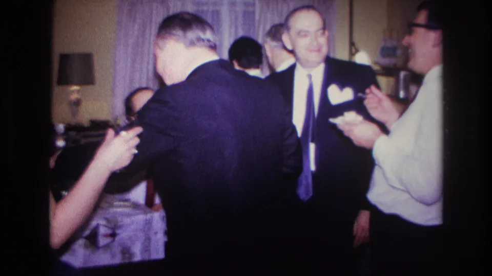 Rockaway Beach, New York, 1967 home-movie frame showing a suited man smiling beside guests eating in a private interior.