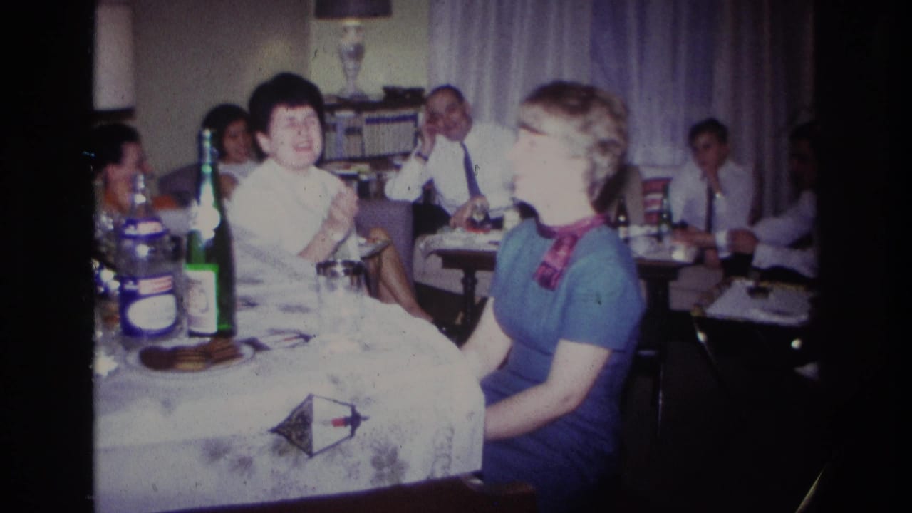 Adults sit at a table with bottles and glasses while talking in a living-room setting.