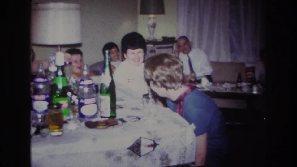 1967 Rockaway Beach home-movie frame with a child and adults at a dining table during a family gathering.