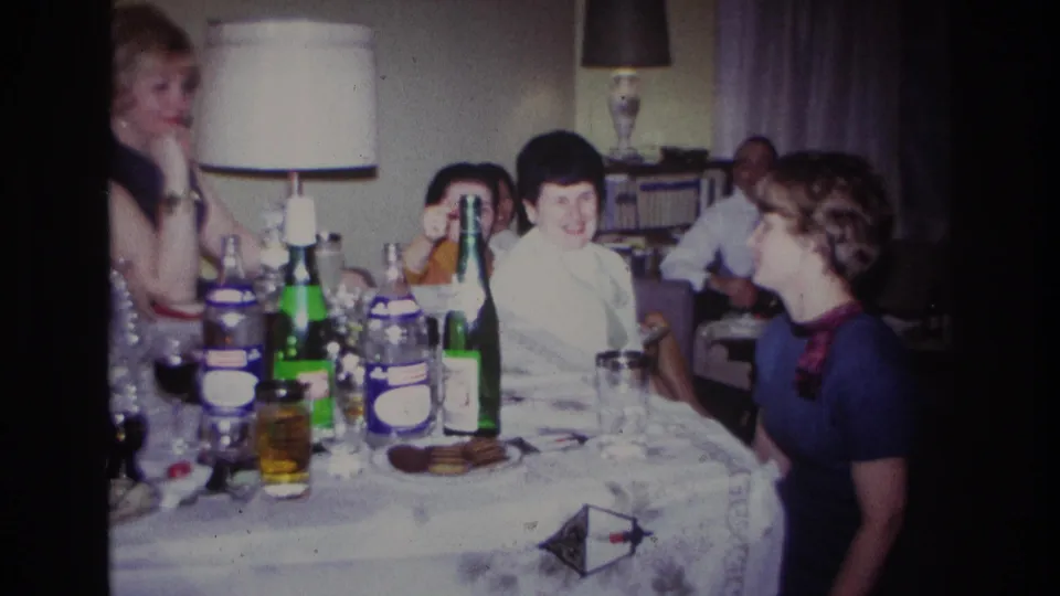 1967 Rockaway Beach frame with several adults seated at a table, including women in light and dark dresses near bottles and glasses.