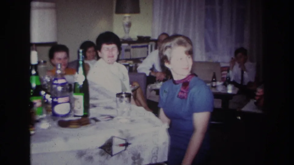 1967 Rockaway Beach home-movie frame showing a woman in a blue dress with a red scarf seated beside bottles, glasses, and other guests.