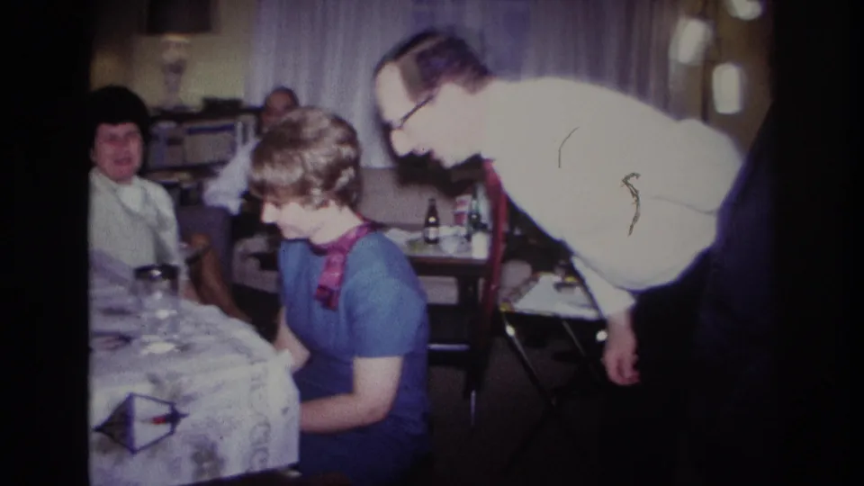 1967 Rockaway Beach frame showing an older man in a white shirt leaning in to speak with a seated woman in a blue dress and red scarf.