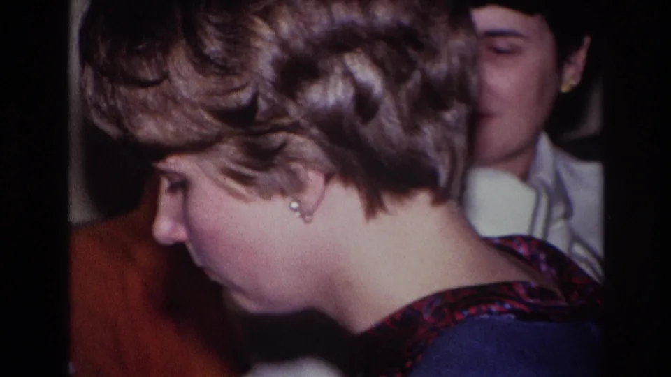 Side view of the blonde woman playing piano, with observers partly visible in the 1967 Rockaway Beach living-room scene.