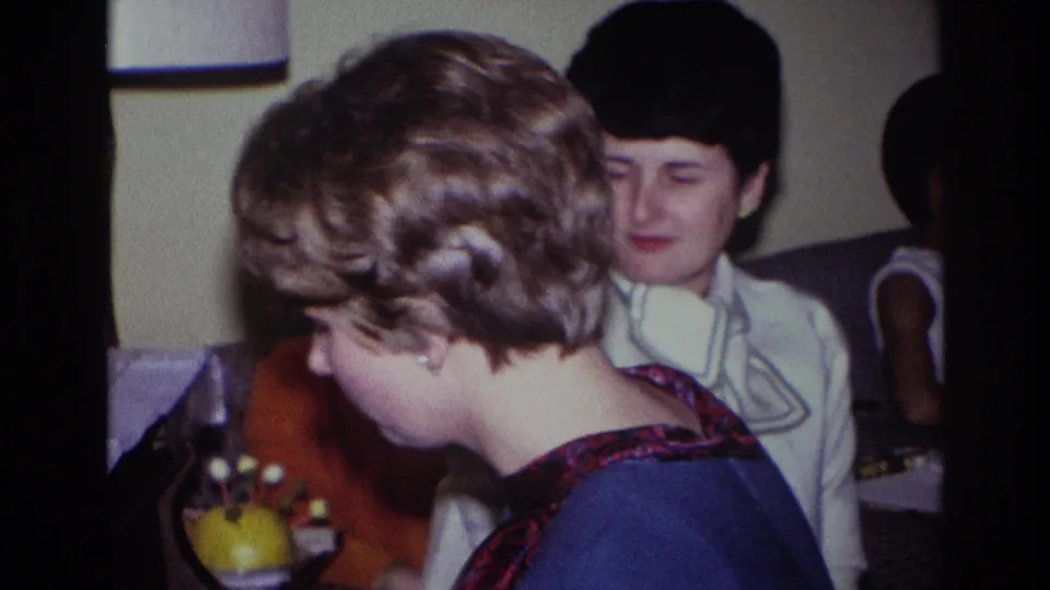 Home-movie frame of the piano player leaning toward the keys, with a seated woman in a white collared shirt visible behind her.