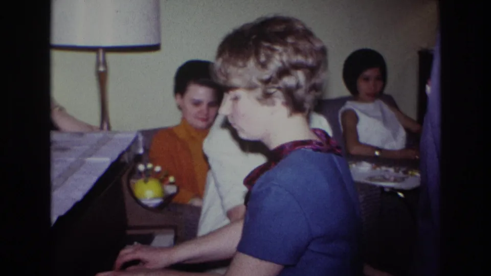 Wider 1967 Rockaway Beach living-room view with the piano player in the foreground and women seated near a lamp and fruit bowl.