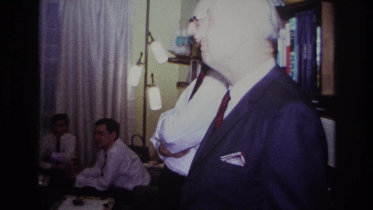 1967 Rockaway Beach home-movie frame showing an older man in a dark suit near seated men inside a private living room.