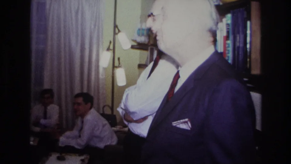 1967 Rockaway Beach home-movie frame showing an older man in a dark suit near seated men inside a private living room.