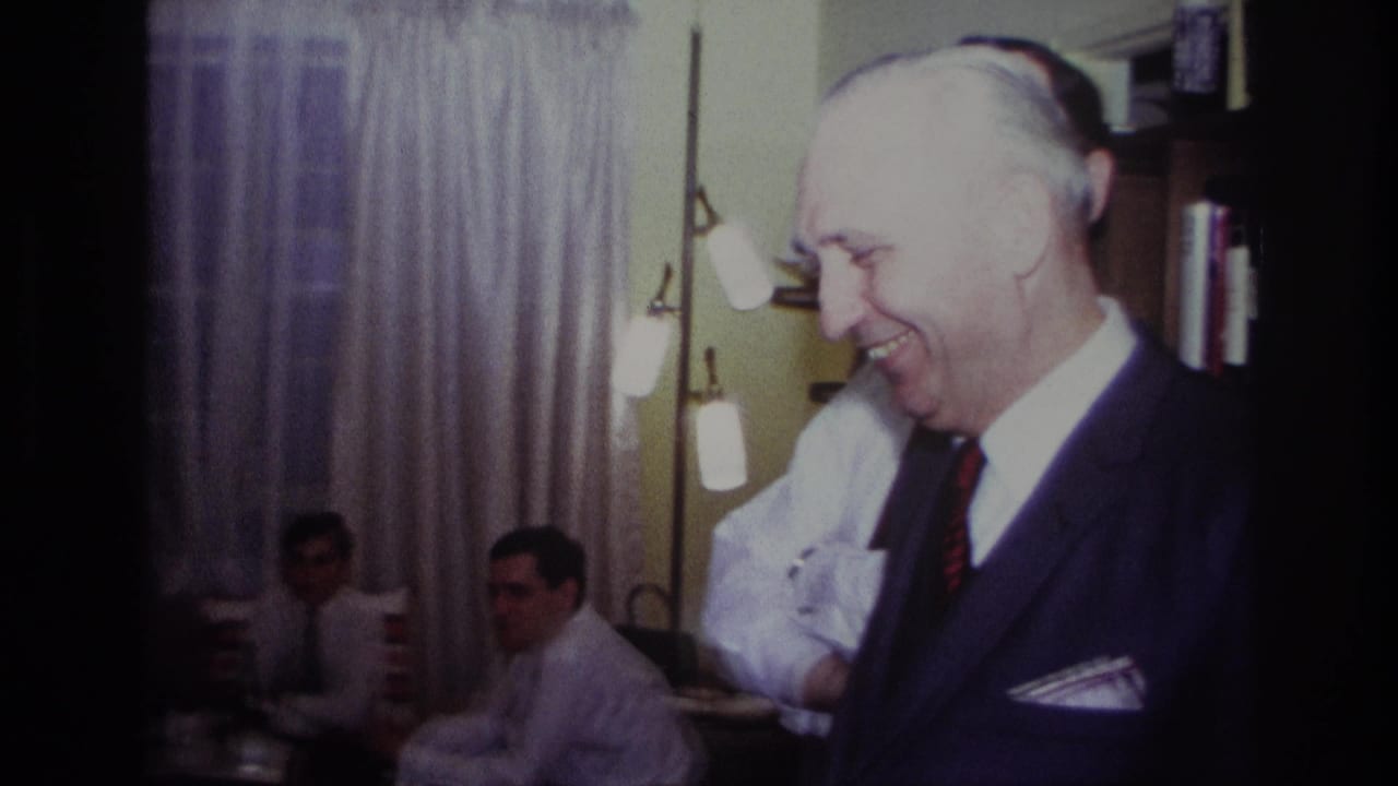 Smiling older man in a suit stands near another man while two seated men sit by curtains.