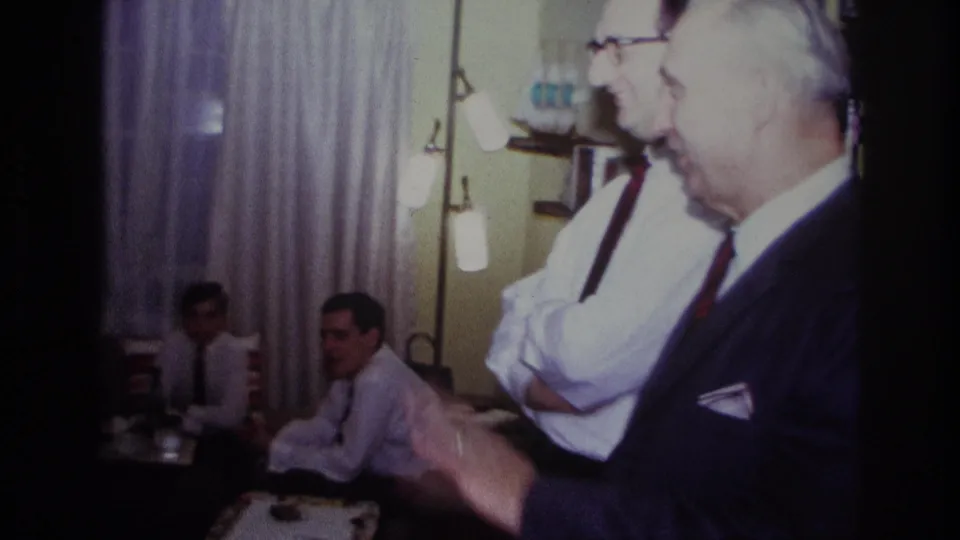 1967 Rockaway Beach home-movie frame showing two standing men in dress clothes and seated guests in a private living room.