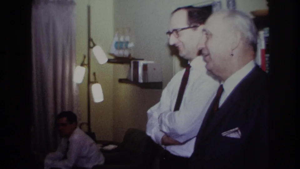 1967 Rockaway Beach frame with two adult men standing in profile and smiling inside a softly lit living room.