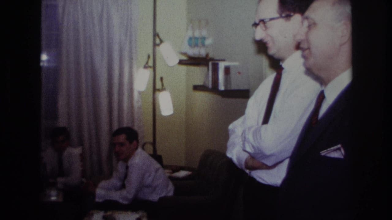 1967 Rockaway Beach home-movie frame showing adults in a living room near a piano, with men in dress shirts and ties in the foreground.