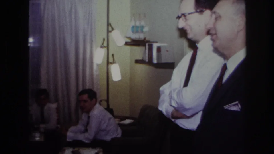 1967 Rockaway Beach home-movie frame showing adults in a living room near a piano, with men in dress shirts and ties in the foreground.