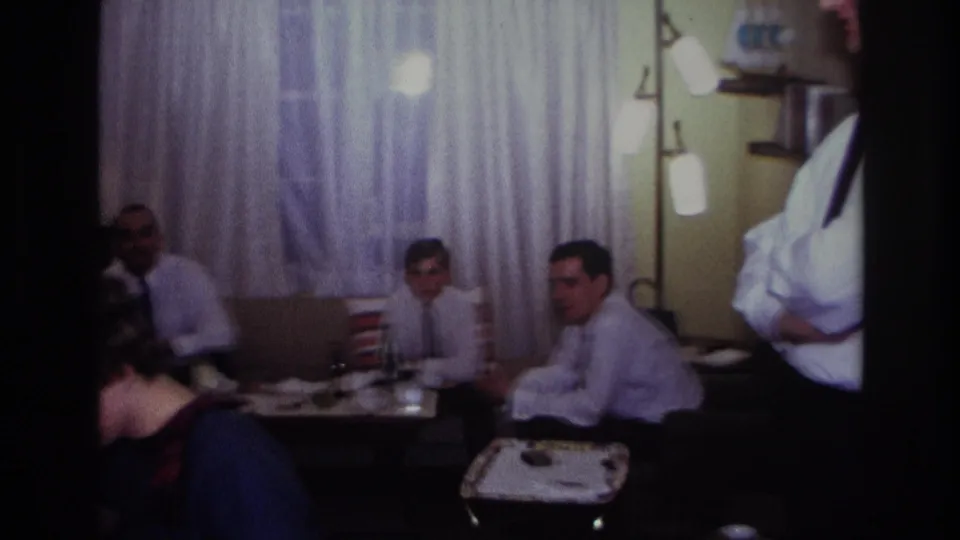 1967 Rockaway Beach living room scene with seated guests, men in shirts and ties, drinks on tables, and sheer curtains behind them.