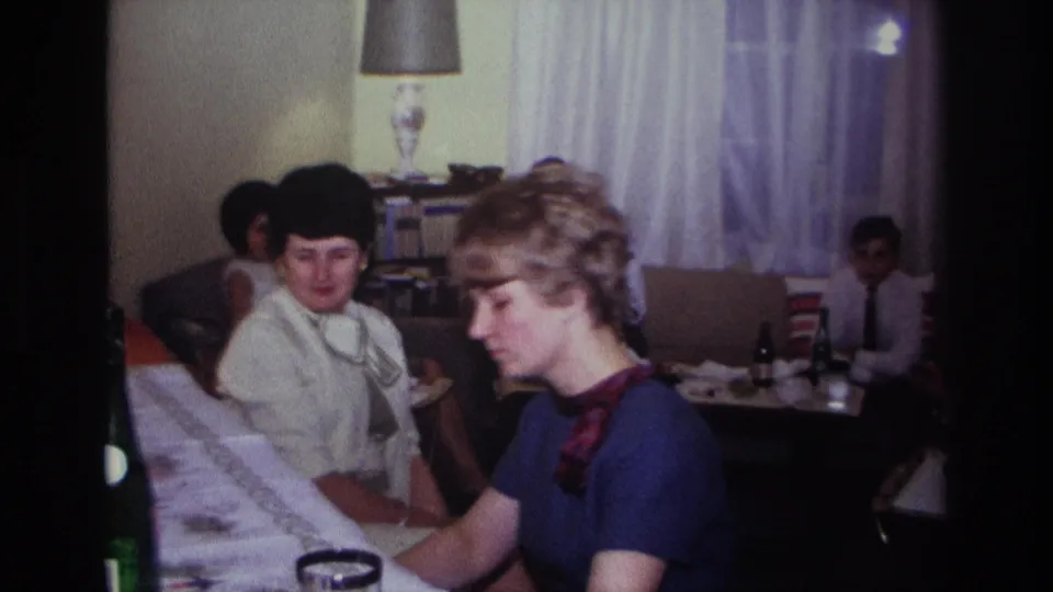1967 Rockaway Beach home-movie frame of two women at a piano, with guests, drink bottles, a lamp, and curtains in the living room.