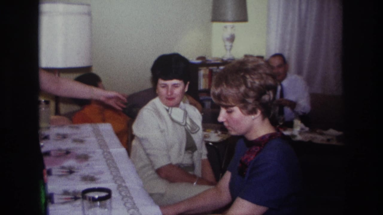 1967 home-movie frame from Rockaway Beach showing piano playing, seated adults, and a gesture across the living room.