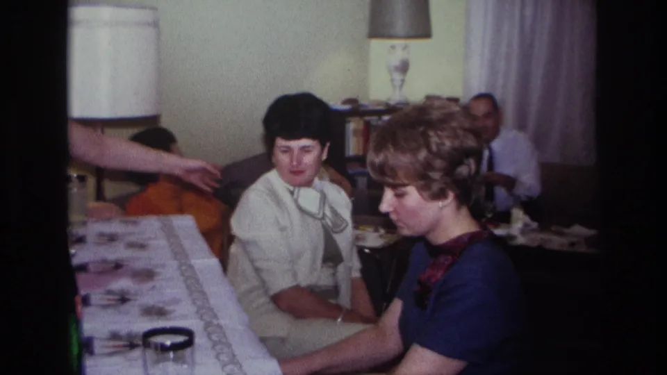 1967 home-movie frame from Rockaway Beach showing piano playing, seated adults, and a gesture across the living room.