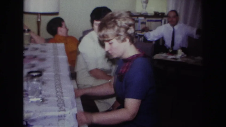 1967 Rockaway Beach living-room frame showing a pianist in blue with family members behind her, including a gesturing man.