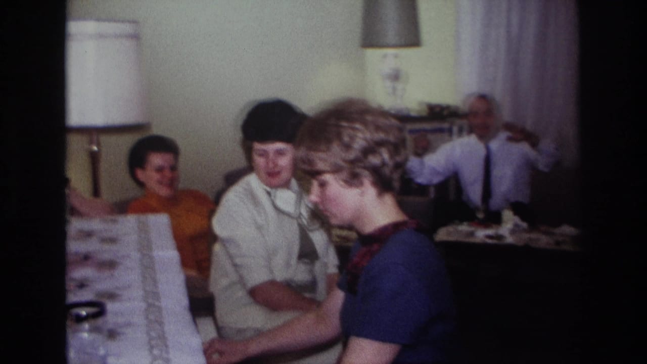 1967 Rockaway Beach home-movie frame showing a woman at a piano as adults sit nearby in a private living room.