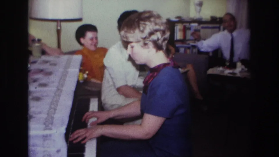 1967 Rockaway Beach home-movie frame showing the woman at the piano, with a patterned cloth and seated adults nearby.