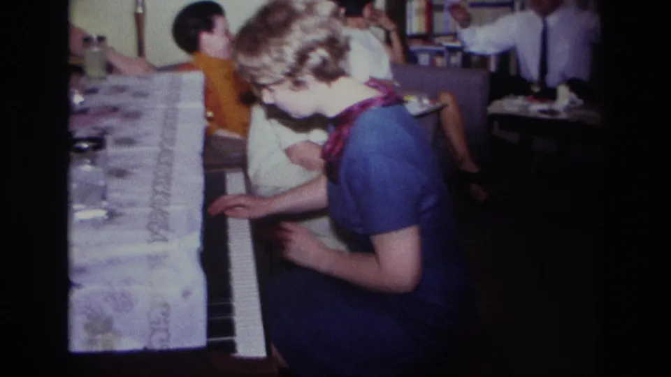 1967 Rockaway Beach frame of a woman seated at the piano, with adults, table items, and shelves visible behind her.